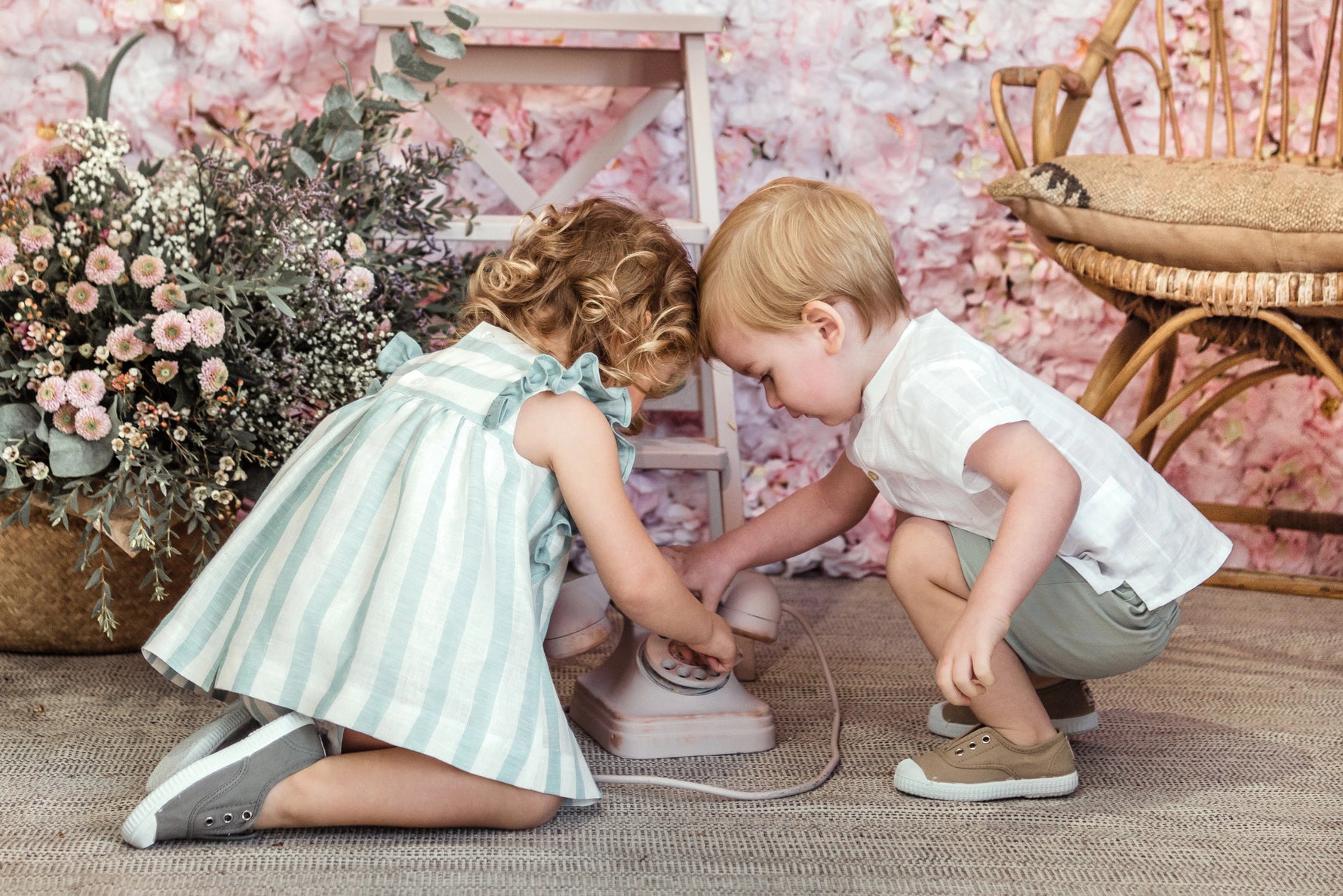 Two children playing with a toy iron in a decorative room with flowers and furniture.