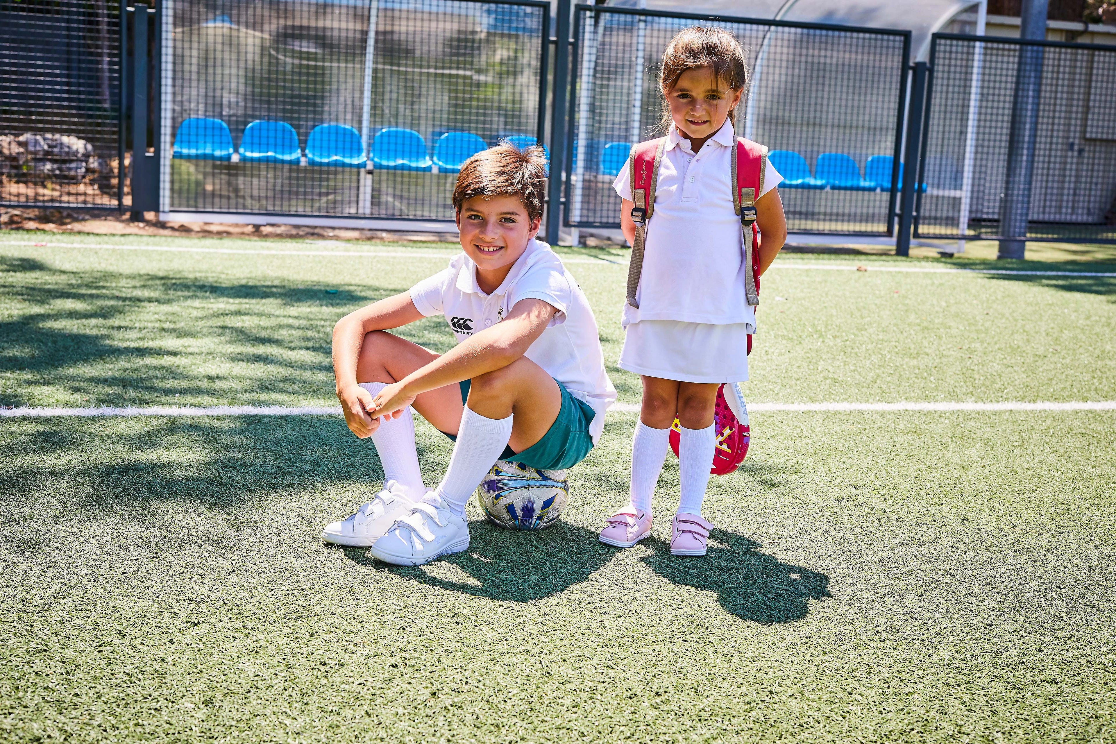 Two children on a sports court, one crouching and the other standing, with sports equipment around.