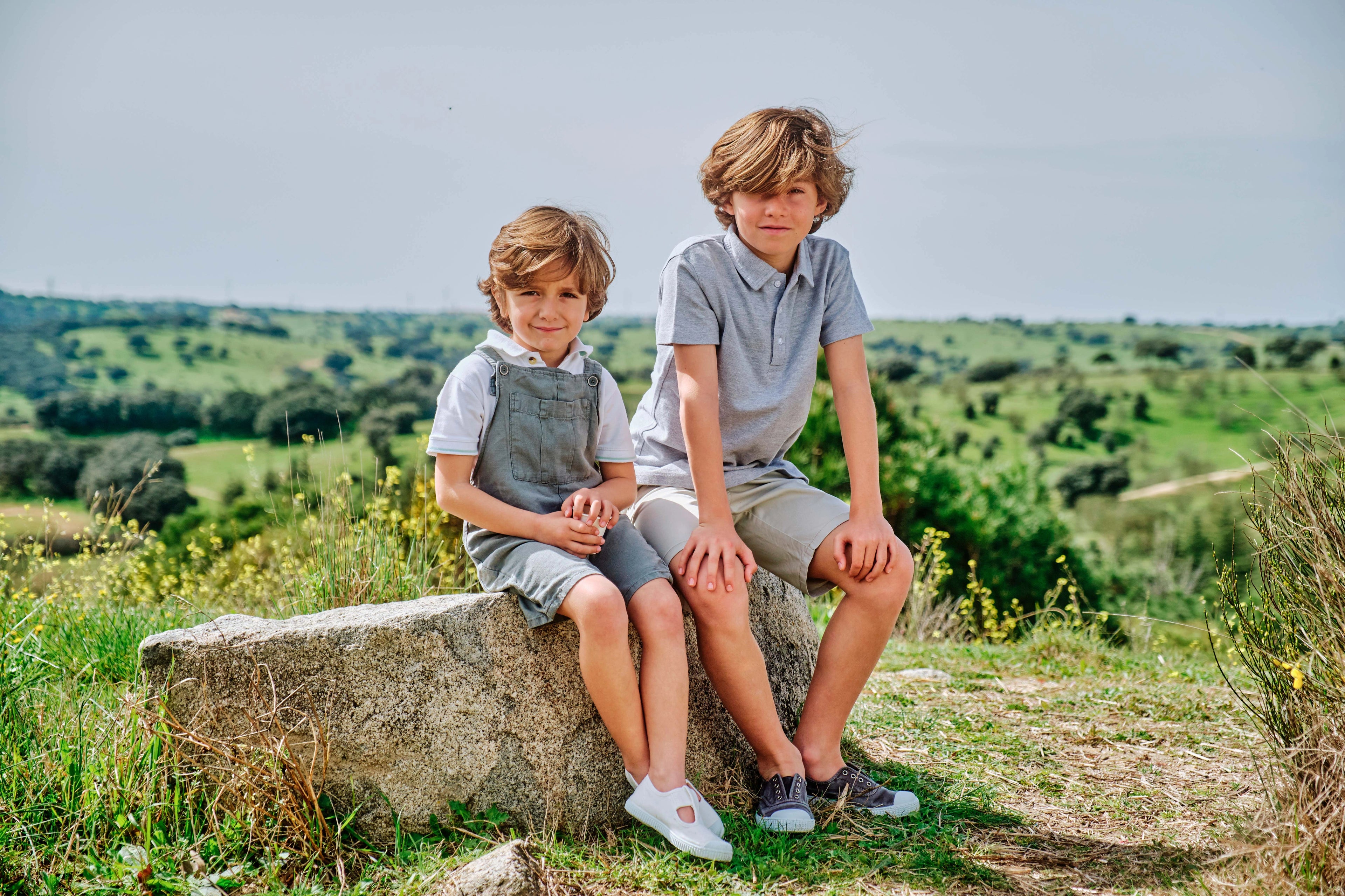 Two children sitting on a rock in a scenic outdoor setting with greenery and a clear sky.