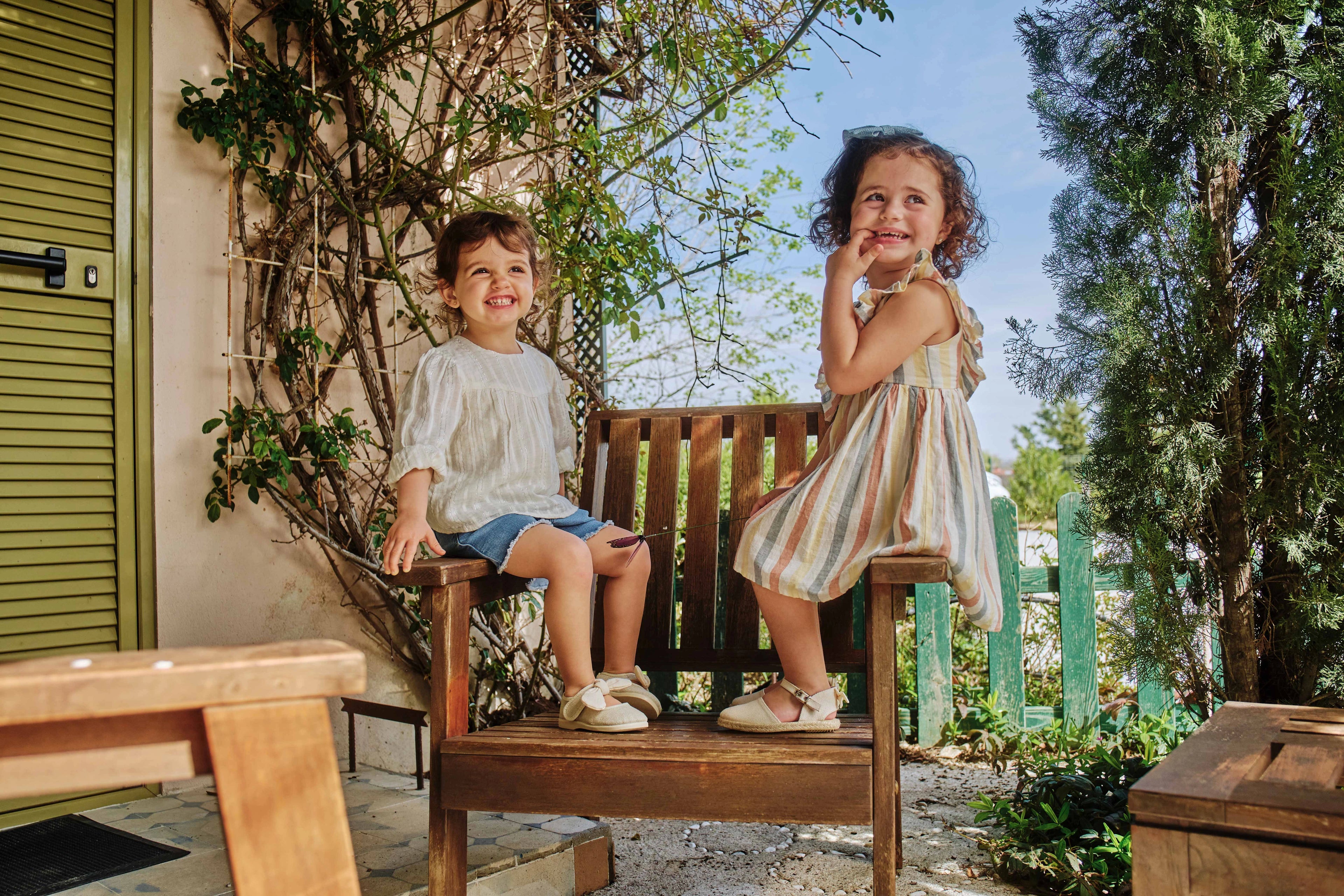 Two children sitting on a wooden bench outdoors with greenery around.